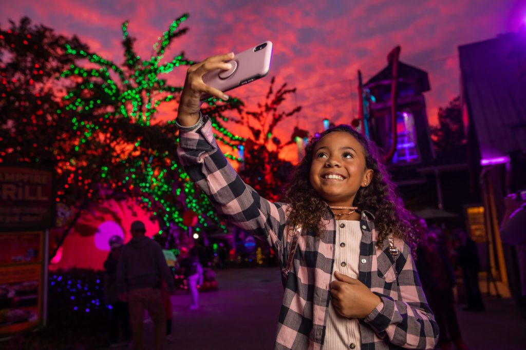 A girl with curly brown hair takes a selfie on a phone at night at the Halloween at Stone Mountain 2019 event.