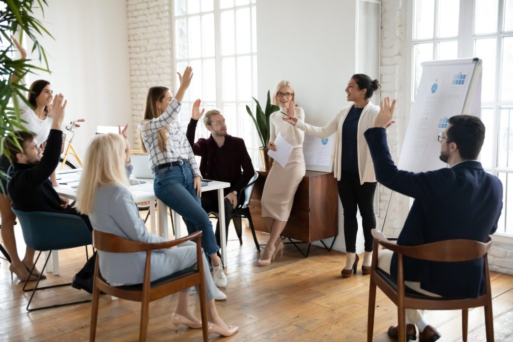 A group of people in business clothes sit and stand together in a meeting as one of them presents charts on an easel. All of them are raising their hands as if asking questions or voting on a question.