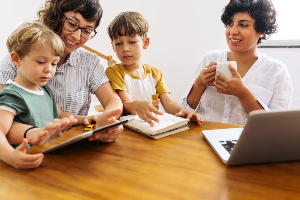 A family sits together at a wooden table. One mom holds a coffee cup and smiles at the other, who is holding a tablet for one child as the other looks over her shoulder and draws in a notebook.