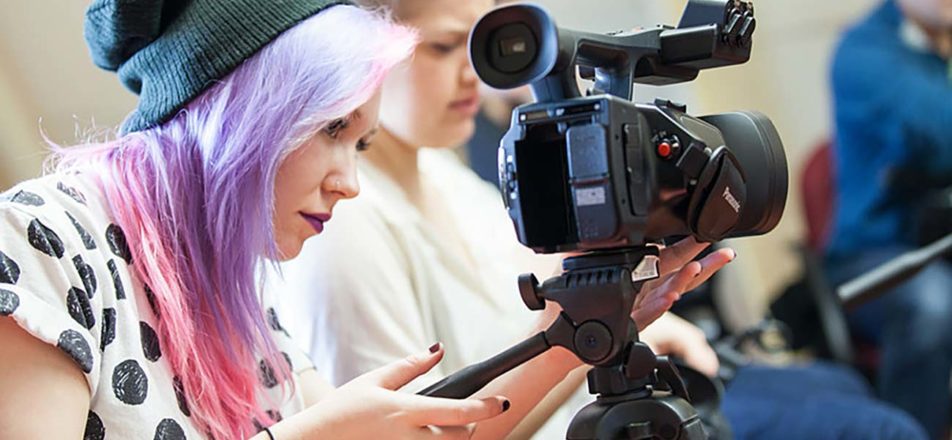 A person with a gray beanie hat and pastel pink-and-purple hair adjusts a movie camera as other people look on in the background.
