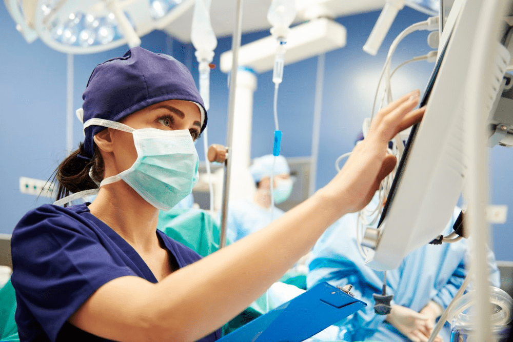 A healthcare worker in a blue surgical mask holds a plastic clipboard and enters information on a touch screen.
