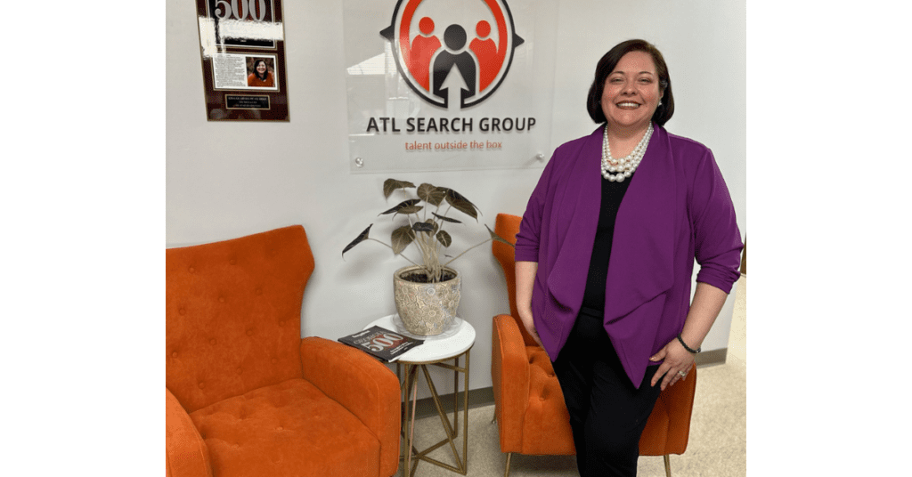 Lisa Guadalupe Clarke stands next to the ATL Search Group sign, in a white office with orange chairs.