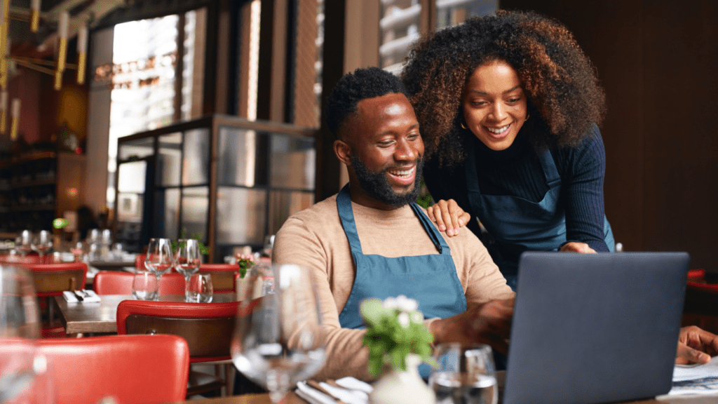 A smiling Black couple look at a laptop together in an empty restaurant that has been prepared for opening.