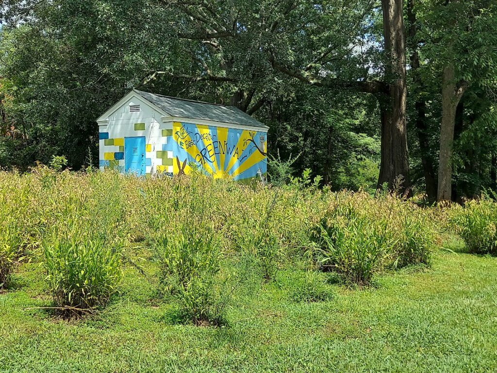 A colorful shed with a rising sun painted on its side, surrounded by green trees and plants. Photo by East Decatur Greenway.