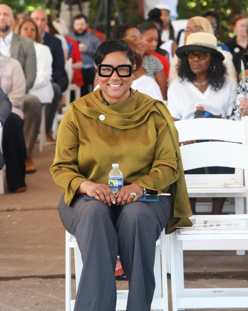 A woman in an elegant gold draped blouse sits in a white folding chair outside, smiling at the camera, with other people sitting in chairs behind her.