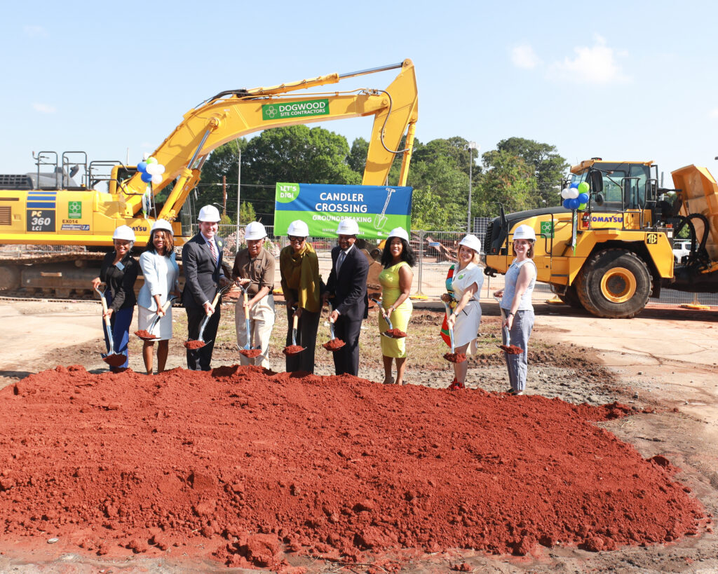 A group of professionally-dressed people in white hard hats hold up shovels with scoops of red dirt in them at a groundbreaking.