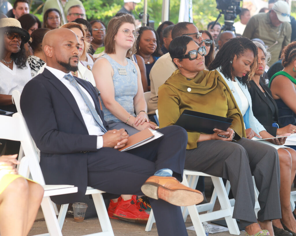 People sit in white folding chairs at an outdoor event and listen attentively.