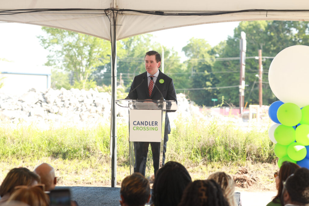 A man in a suit speaks into microphones at a transparent podium with a "Candler Crossing" sign on the front.