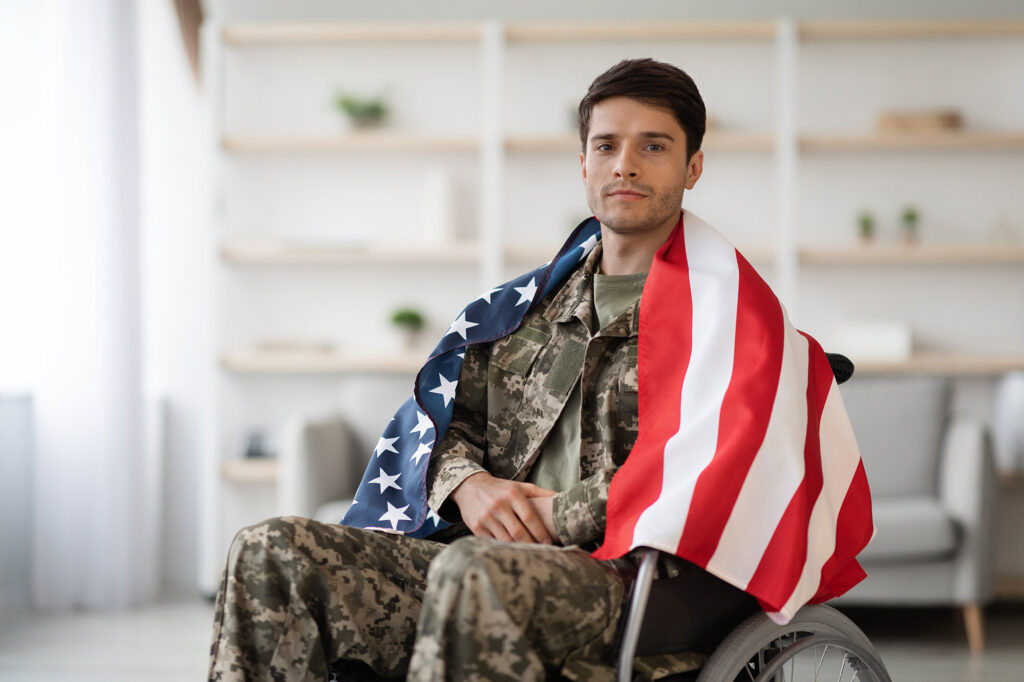 Handsome young military man in uniform sitting in wheelchair with American flag draped over his shoulders.