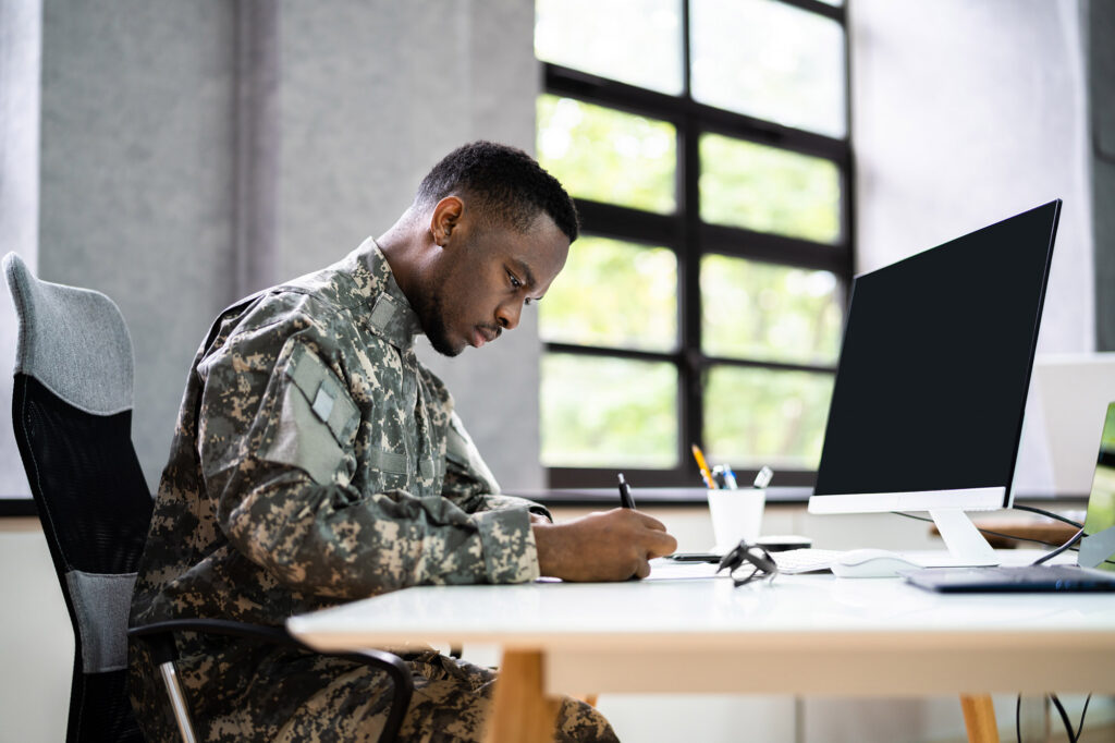 African-American soldier sitting at a desk with a computer, writing on a piece of paper with a focused look on his face.